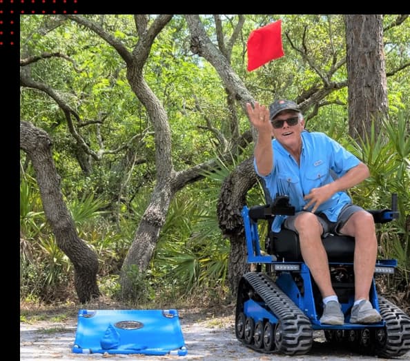 A rider in a blue EcoRover tossing a red bean bag in a wooded outdoor setting.