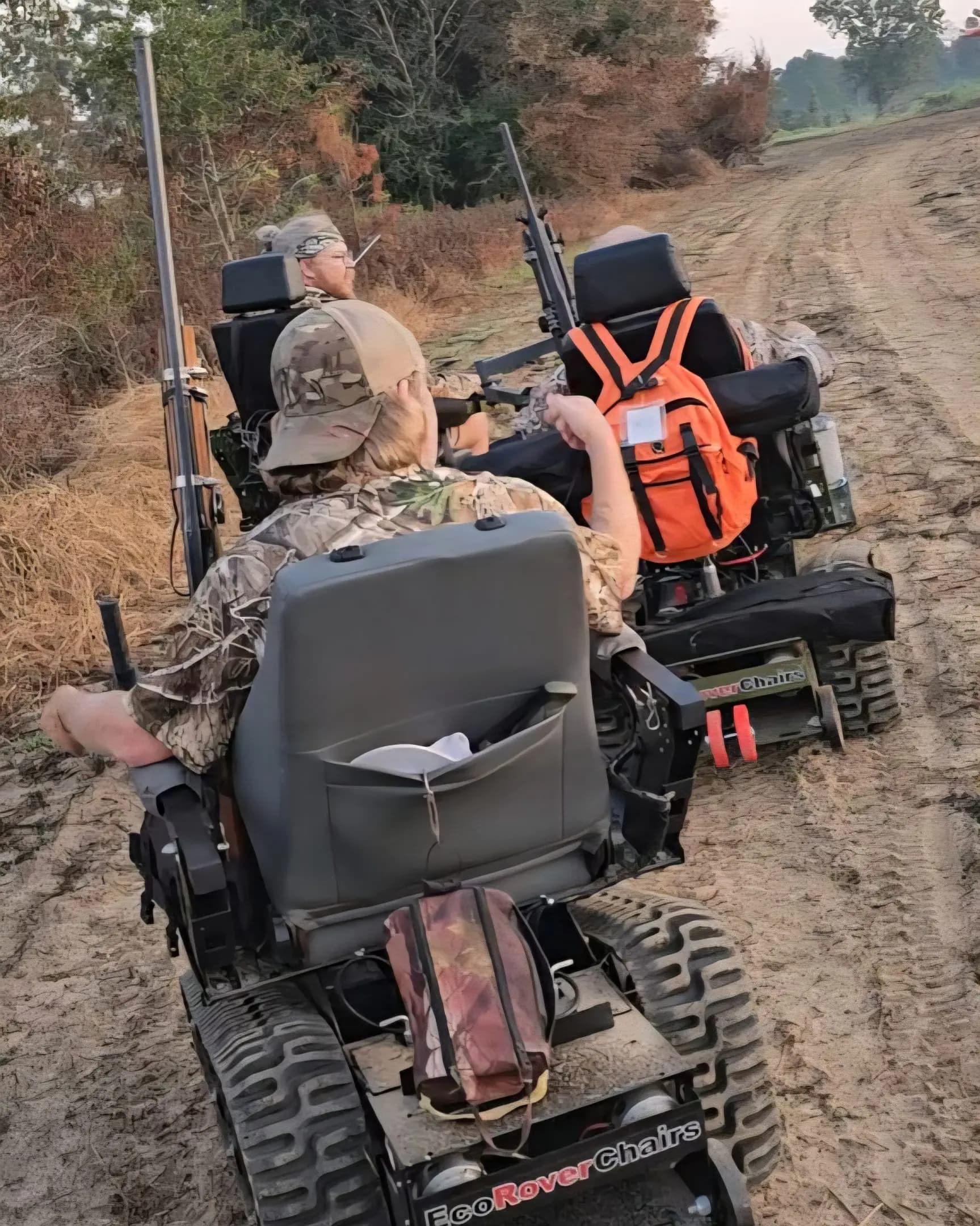Two hunters in EcoRovers heading down a dirt road with rifles and gear, seen from behind.